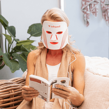 Woman using the Infraredi LED Red Light Therapy Mask while reading a book in a cozy setting.