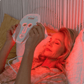 Woman using Infraredi LED Red Light Therapy Mask for skincare in bed, promoting youthful and glowing skin with red light therapy.